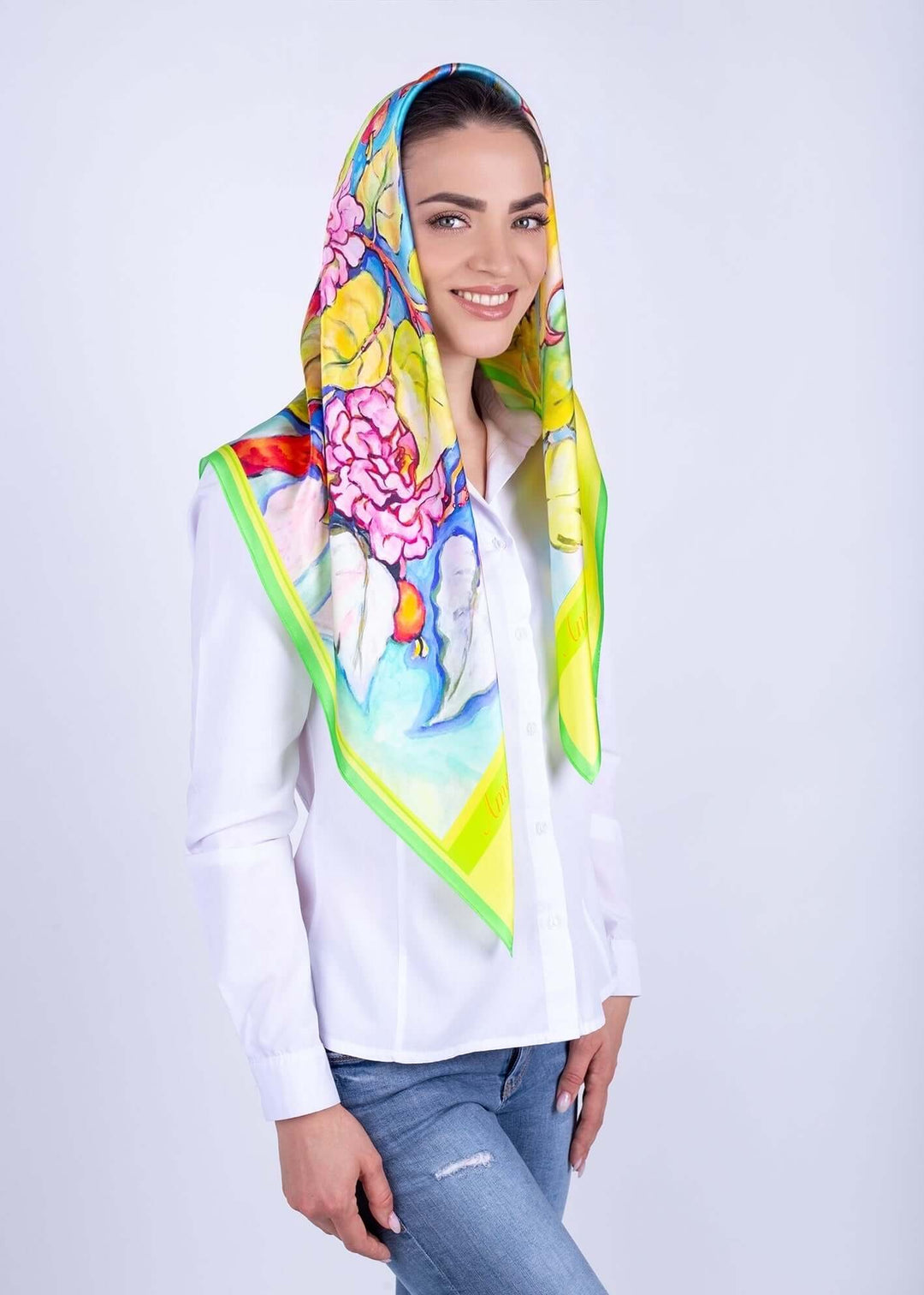Frau mit buntem Seidenschal in Gelb und Blumenmotiv, lächelnd, in weißem Hemd und blauen Jeans, vor neutralem Hintergrund.