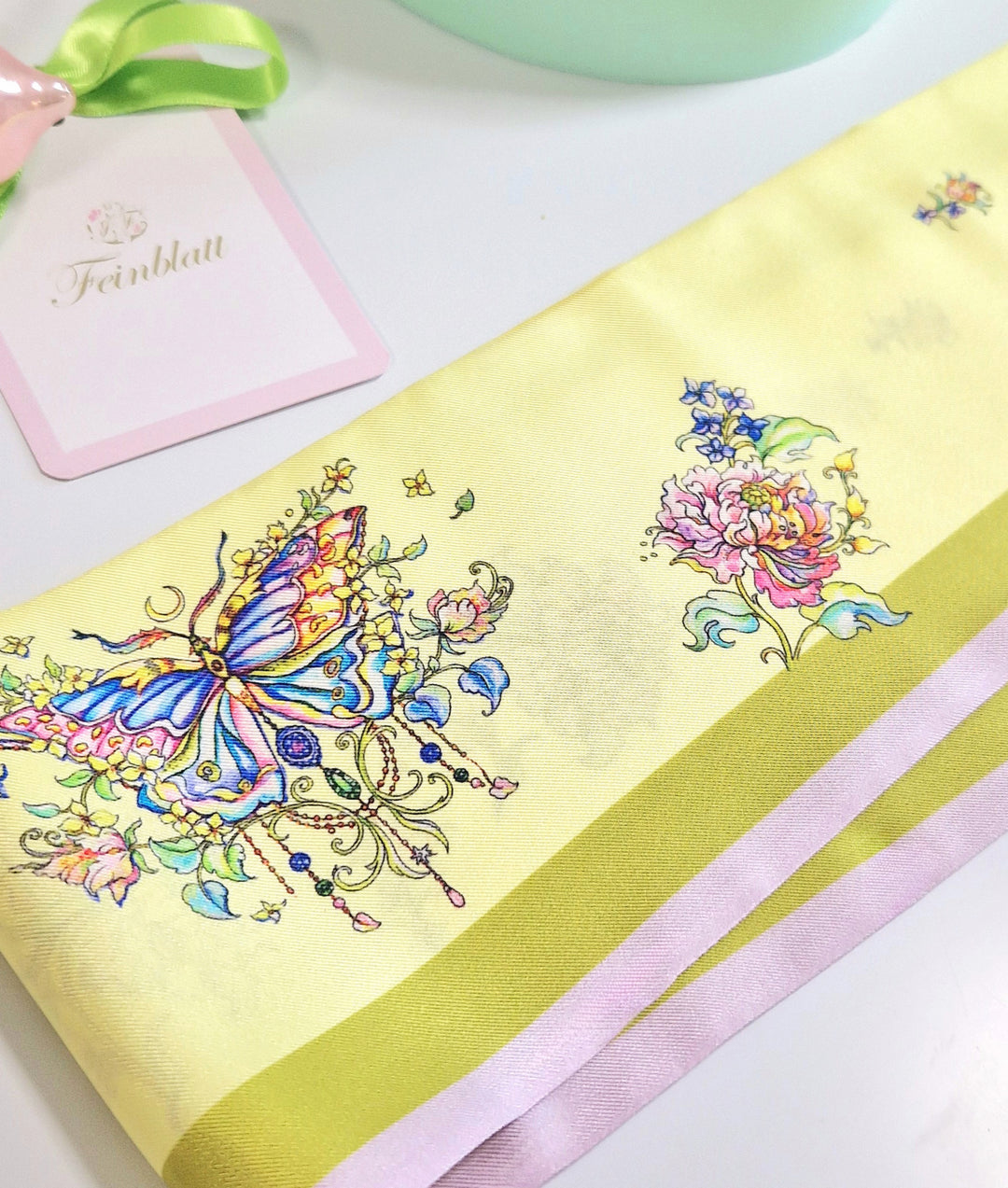 Detailaufnahme eines gefalteten gelben Seidenschals mit Schmetterling, Pfingstrose und Schmuckmotiven, daneben ein Feinblatt-Anhänger.
