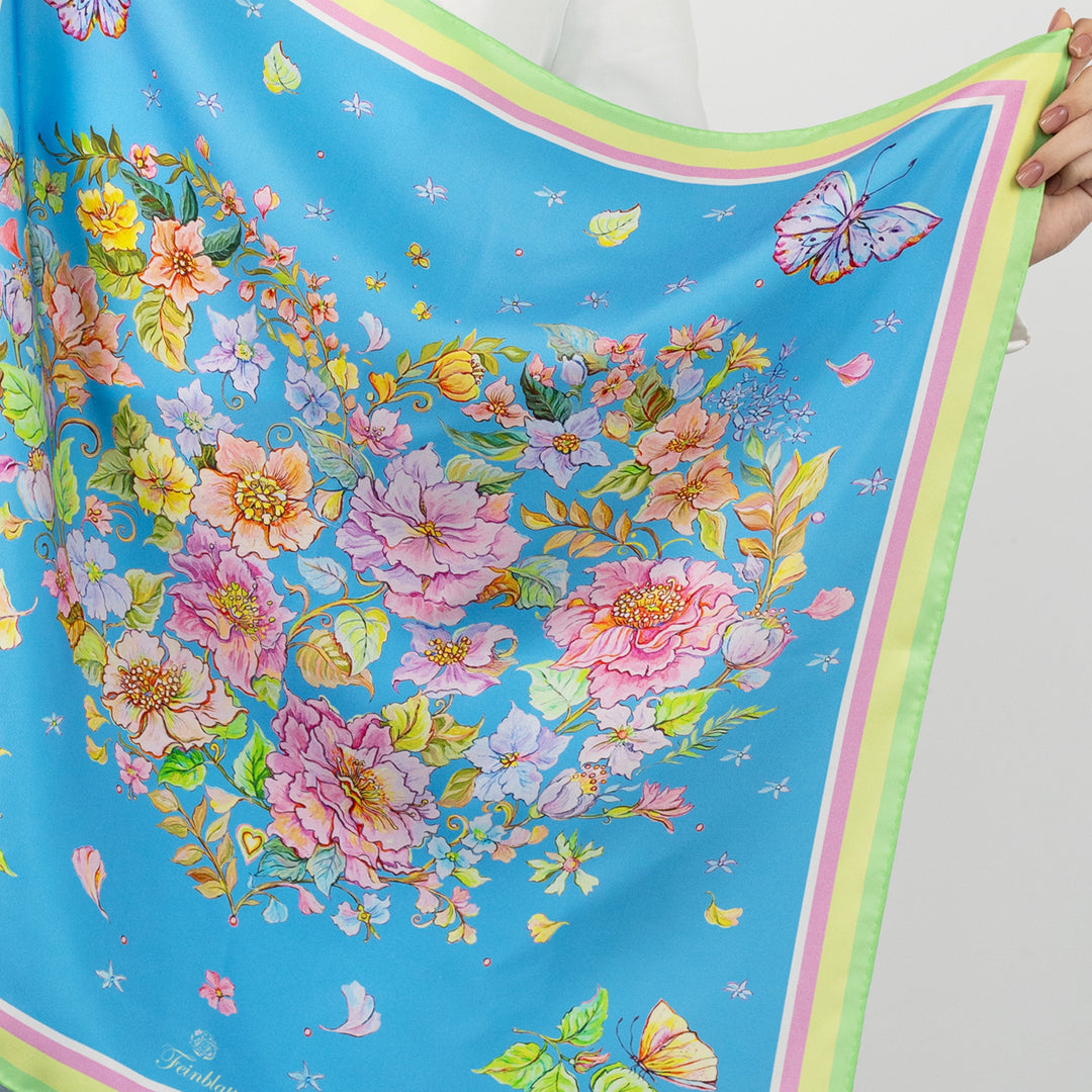 Detailansicht eines blauen Seidentuchs von Feinblatt mit floralem Herzmotiv, Schmetterlingen und grün-gelb-rosa Bordüre, von einer Frau gehalten.