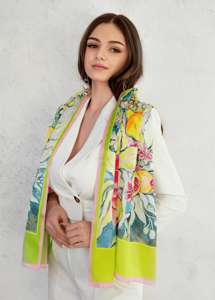Ein leichter Seidenschal mit einem leuchtenden Obst und Blumenmotiv in Gelb, Rosa und Grün, locker über die Schultern einer Frau in einem weißen Kostüm geworfen – ein exklusives Sommeraccessoire von Designerin Iryna Feinblatt