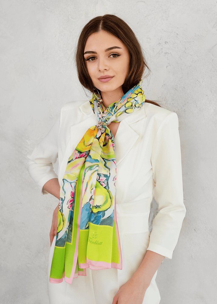 Ein modisches Seidenhalstuch mit einem farbenfrohen Blumen- und Fruchtmotiv in den Farben Gelb, Grün und Pink, das elegant um den Hals einer Frau im weißen Hosenanzug gebunden ist – ein stilvolles Designeraccessoire von Iryna Feinblatt