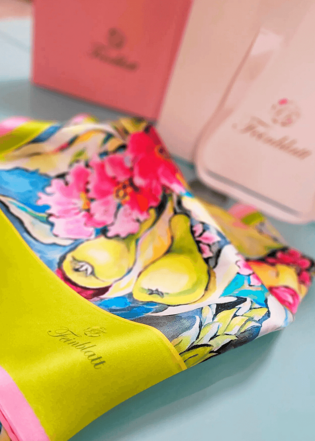 Dieser aufwendig gearbeitete Seidenschal mit einem auffälligen Motiv aus exotischen Sommerfrüchten und Blumen in Grün, Rosa und Blau auf glänzendem Seidensatin mit „Feinblatt“-Logo wird in einer stilvollen Geschenkverpackung präsentiert und ist somit das ideale edle Geschenk.