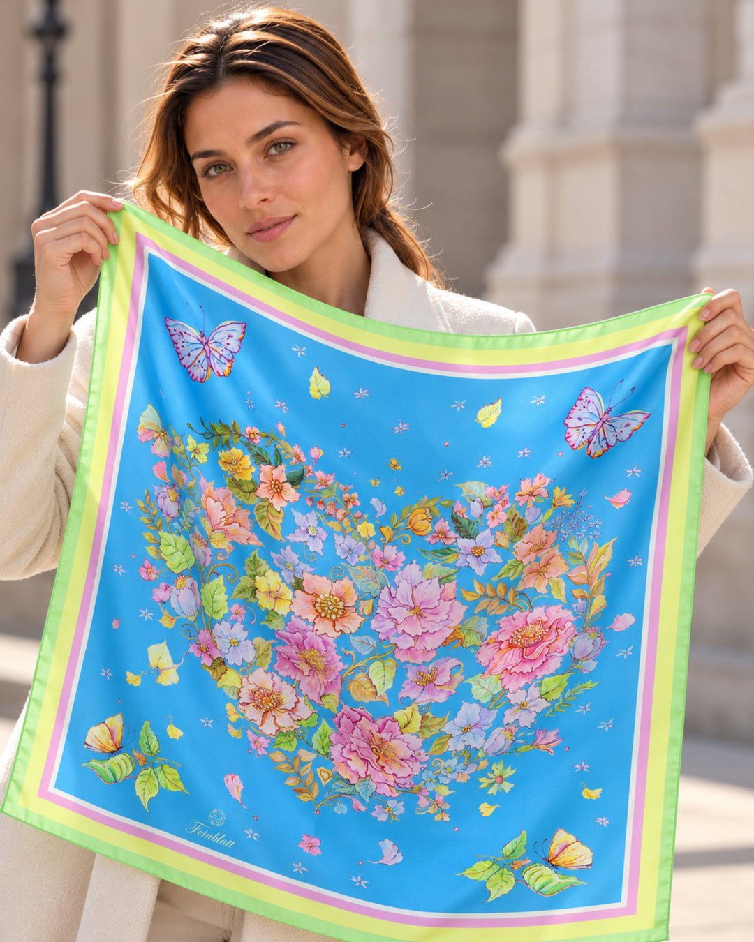 Frau präsentiert einen blauen Designer-Seidenschal von Feinblatt mit floralem Herzmotiv, Schmetterlingen und grüner Bordüre.