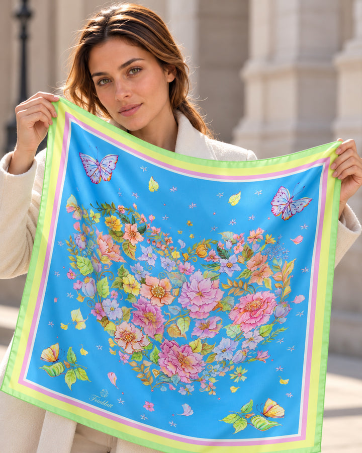 Frau präsentiert einen blauen Designer-Seidenschal von Feinblatt mit floralem Herzmotiv, Schmetterlingen und grüner Bordüre.