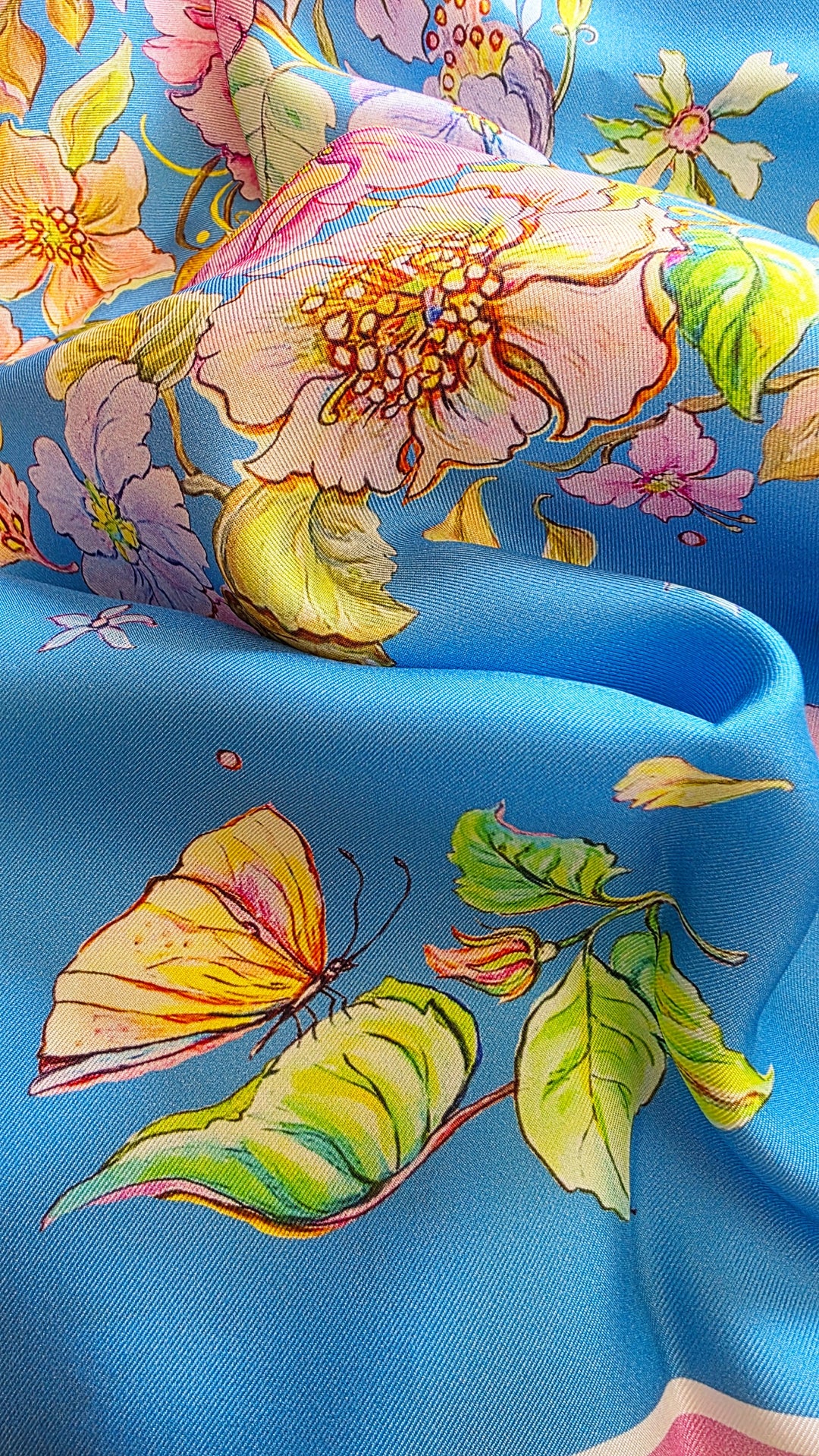Nahaufnahme eines blauen Feinblatt Seidenschals mit gelbem Schmetterling, grünen Blättern und zarten Blüten auf fein gewebter Seide.