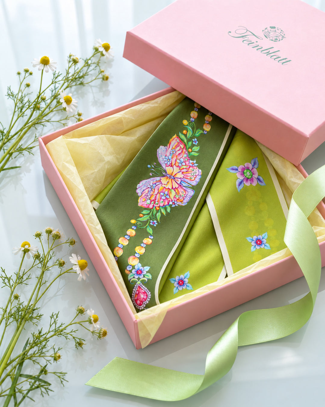 Olivgrüner Seidenschal mit buntem Schmetterlingsmotiv, in einer rosa Feinblatt Geschenkbox mit hellgelbem Seidenpapier und kleinen Kamillenblüten.